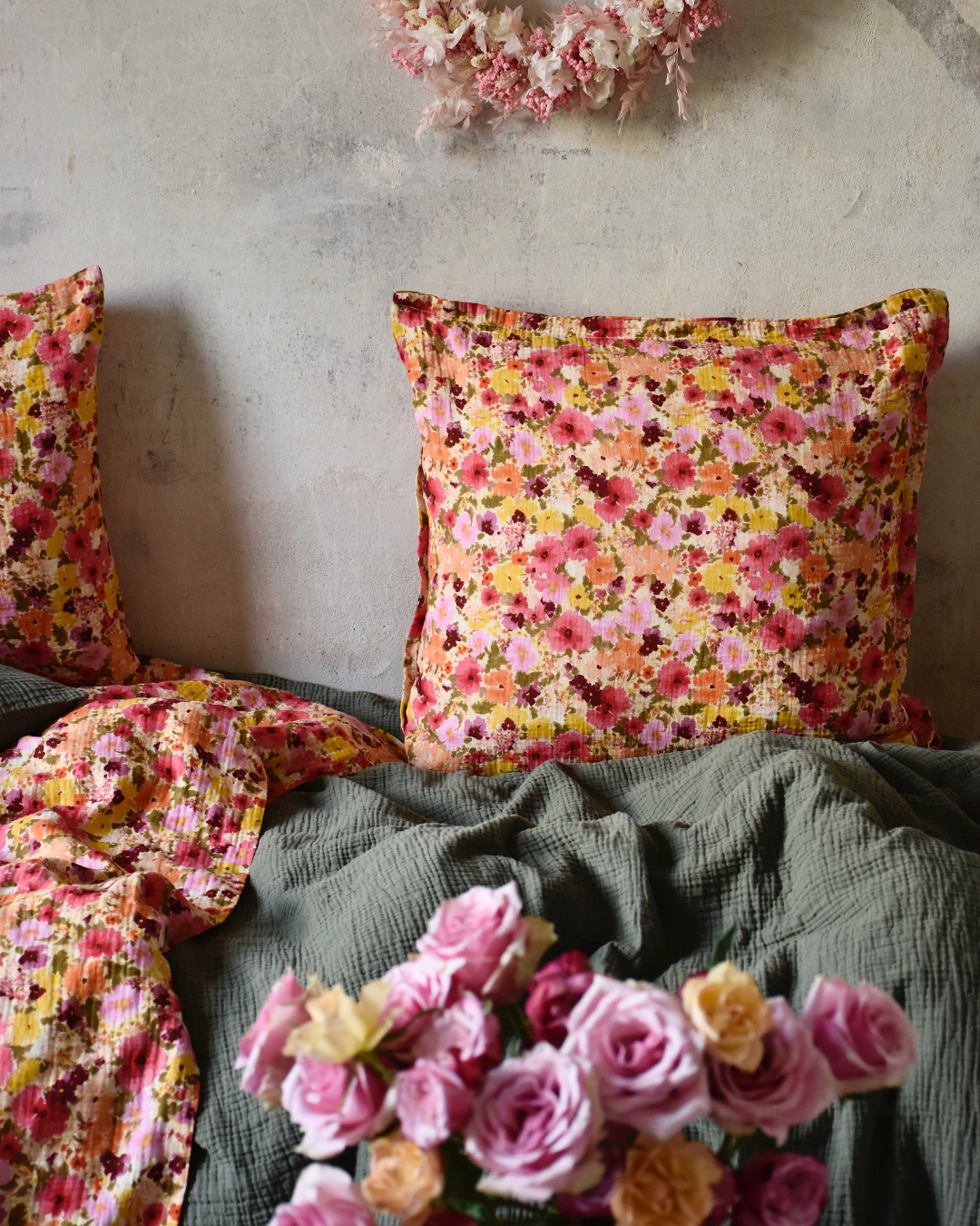 Kissen mit buntem Blumenmuster auf olivgrüner Musselin-Bettwäsche vor einer hellen Wand